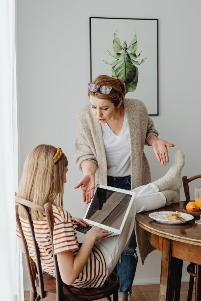 A mother expresses displeasure as her teenage daughter uses a laptop during breakfast.